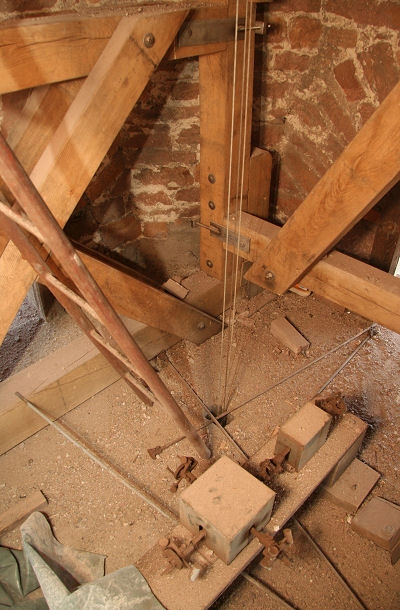 Nun noch ein kurzer Blick in den Turm.
Die eigentliche Uhr befindet sich ein ganzes St&uuml;ck unterhalb des Glockenstuhls.
Die Zeiger werden durch eine Welle angetrieben die von der Uhr aus senkrecht nach oben durch die Decke der Glockenstube f&uuml;hrt.
Dort wird die Drehbewegung durch ein Getriebe in dem kleinen K&auml;stchen in der Mitte auf 4 weitere Wellen umgesetzt. Diese treiben die Zeiger der 4 Ziffernbl&auml;tter an. Die Untersetzung 60 : 1 zwischen Stunden- und Minutenzeiger erfolgt separat in 4 weiteren Getrieben direkt hinter den Ziffernbl&auml;ttern.

Neben dem Verteilerk&auml;stchen sehen Drahtseile durch ein Loch im Boden laufen.
&Uuml;ber diese bet&auml;tigt die Uhr die H&auml;mmer f&uuml;r den Glockenschlag.
